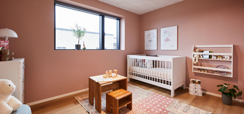 Hyggeligt børneværelse med rosa vægge, hvid tremmeseng, lille bord, væghængt bogreol, stort vindue og mønstret gulvtæppe – inspirerende indretning til nybyg og familiens husdrømme.