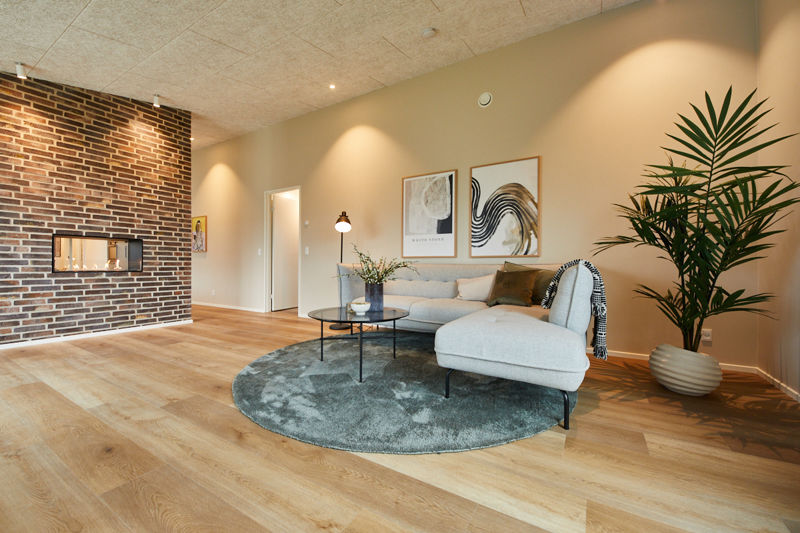Stue fra eurodan-huse med lys sofa og chaiselong, rundt grønt tæppe, sort sofabord, stor plante, murstensvæg med pejseindsats og varme spotlys – inspiration til stemningsfuldt nybyggeri, husbyggeri og moderne husdrømme.
