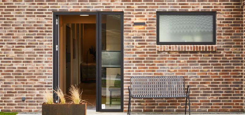 Indgangsparti fra eurodan-huse med rødbrun murstensfacade, sort hoveddør med højt sideparti, smalt vandret vindue, udendørslampe, plantekasse og bænk ved muren – inspiration til døre, entréfacade og nybyggeri med karakter.