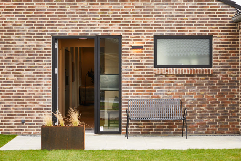 Indgangsparti fra eurodan-huse med rødbrun murstensfacade, sort hoveddør med højt sideparti, smalt vandret vindue, udendørslampe, plantekasse og bænk ved muren – inspiration til døre, entréfacade og nybyggeri med karakter.