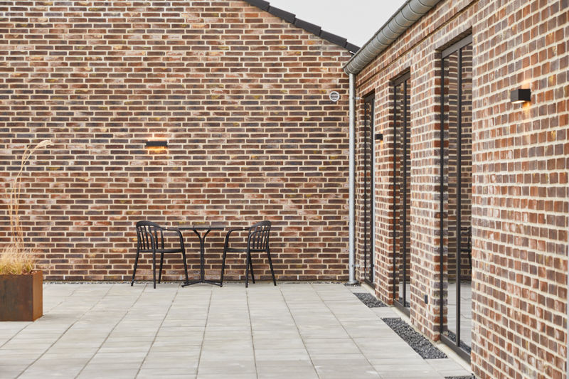 Afskærmet terrasse fra eurodan-huse med rødbrun murstensfacade, sorte glasdøre, lille cafébord med stole, væglamper, grå fliser og plantekasse med prydgræs – inspiration til nybygget hus med intim terrasse og moderne uderum.