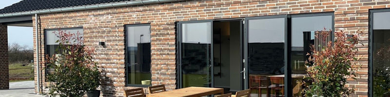 Murstensfacade fra eurodan-huse med sorte vinduer, åbne terrassedøre, ovenlysvinduer i taget, solceller og terrasse med træbord og stole – inspiration til moderne vinduespartier, døre og energibevidst nybyggeri.