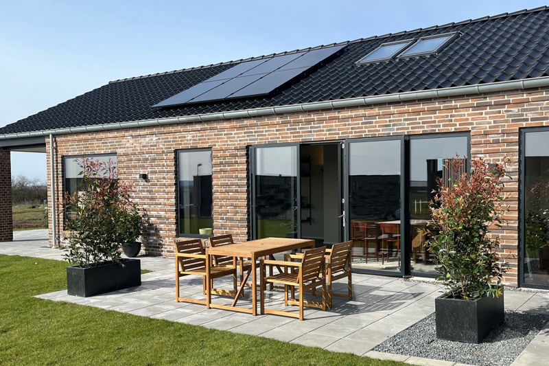 Murstensfacade fra eurodan-huse med sorte vinduer, åbne terrassedøre, ovenlysvinduer i taget, solceller og terrasse med træbord og stole – inspiration til moderne vinduespartier, døre og energibevidst nybyggeri.