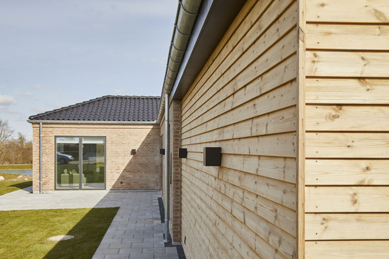 Terrasse og facade fra eurodan-huse med vandret træbeklædning i forgrunden, lys murstensfacade, sorte skydedøre, grå terrassefliser og græsplæne langs huset – inspiration til nybyggeri med trædetaljer, terrasse og moderne husbyggeri.