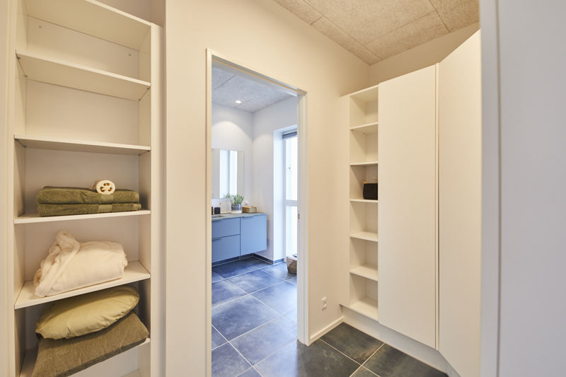 Walk-in garderobe fra eurodan-huse med hvide reoler, åbne hylder med håndklæder og puder, højt skab, mørkt klinkegulv og kig til badeværelse med vaskemøbel og stort spejl – inspiration til praktisk opbevaring tæt på bad i nybyggeri.