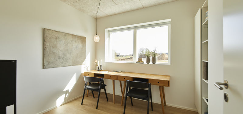 Kontor fra eurodan-huse med langt skrivebord i træ, to sorte stole, hvid reol, stor abstrakt vægkunst, pendellampe og bredt vindue med planter i karmen – inspiration til hjemmearbejde, nybyggeri og funktionelle værelser.