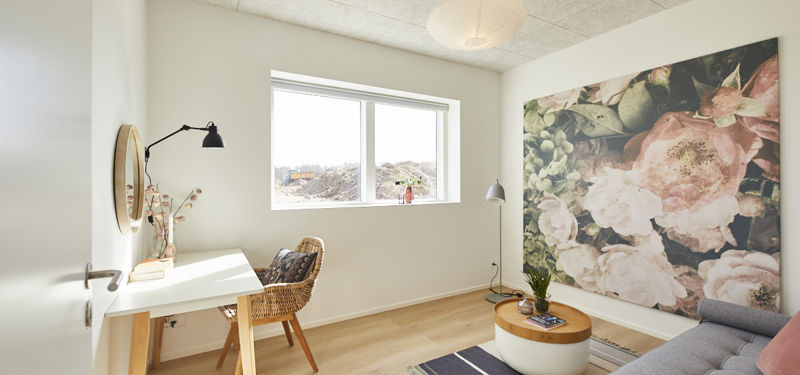Kontor og opholdsrum fra eurodan-huse med hvidt skrivebord, fletstol, rundt spejl, stor blomstret vægdekoration, grå sofa, rundt bord og bredt vindue – inspiration til fleksibelt hjemmekontor, gæsteværelse og nybyggeri.
