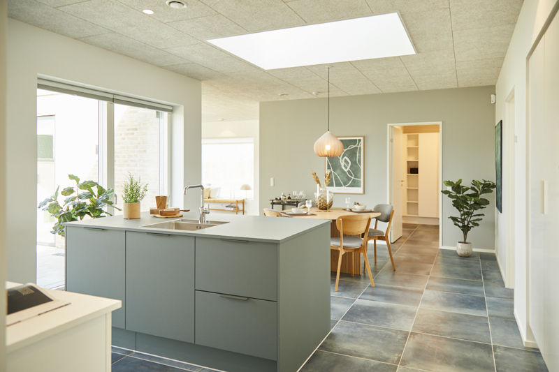 Køkken-alrum fra eurodan-huse med grågrøn køkkenø, vask og armatur, stor firkantet ovenlysflade, mørke klinker på gulvet, spiseplads med rund lampe, grønne planter, bredt vinduesparti og åben dør til depot med hylder – inspiration til nybyggeri, husbyggeri og funktionelle husdrømme.