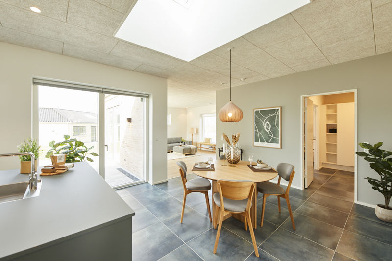 Åbent køkken-alrum fra eurodan-huse med køkkenø i forgrunden, rundt spisebord med træstole, stor pendel over bordet, ovenlys i akustikloft, terrassedør med udsyn, grøn vægkunst og åbent depot/opbevaringsrum ved siden af spisepladsen – inspiration til nybyggeri med gennemtænkt planløsning, familieliv og moderne husdrømme.