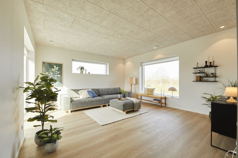 Rummelig stue fra eurodan-huse med stor grå hjørnesofa, matchende puf, stort sansevindue med bænk, højt smalt vindue, væghylder og akustikloft – inspiration til nybyggeri, husbyggeri og funktionelle husdrømme.