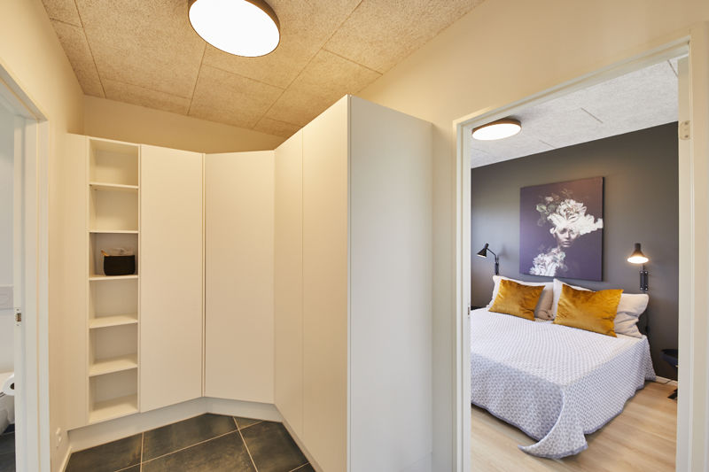 Walk-in closet fra eurodan-huse med hvide skabe, åbne reoler, hjørneskab, mørkt klinkegulv og direkte adgang til soveværelse med mørk væg, sengebordslamper og gule puder – inspiration til funktionel opbevaring i nybyggeri og husdrømme.