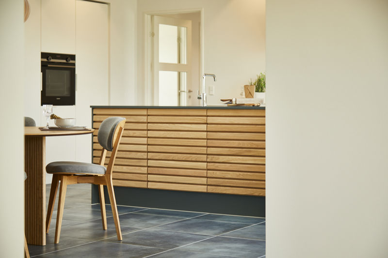 Detalje af køkkenø fra eurodan-huse med vandrette lameller i træ, mørk sokkel, bordplade med armatur og lille plante, spisebordsstol i forgrunden og høj skabsvæg med indbygget ovn i baggrunden – konkret inspiration til køkkenø, opbevaring og materialevalg i moderne nybyggeri og husbyggeri.