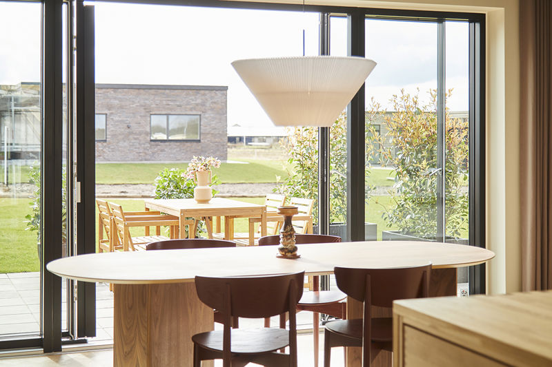 Spiseplads fra eurodan-huse med store sorte skydedørspartier, åben adgang til terrasse, rundt spisebord, træstole, stor hvid pendel og udsyn til haven – inspiration til vinduer og døre i nybyggeri med tæt kontakt til uderummet.