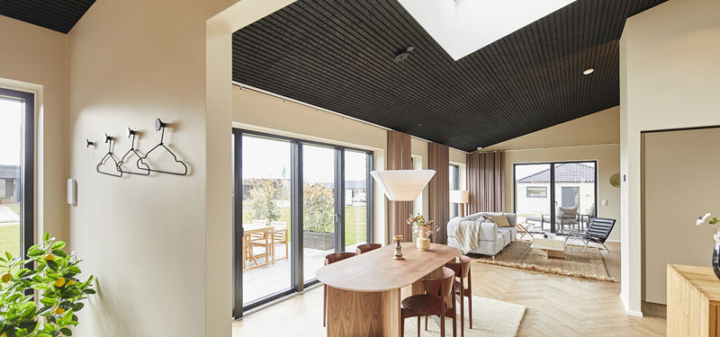Åbent opholdsrum fra eurodan-huse med spisebord i træ, hvide pendler, store vinduespartier, sort akustikloft med ovenlys, lyse gulve og direkte kig fra køkken-alrum til stue med sofa og loungestol – inspiration til nybyggeri med sammenhæng mellem madlavning, spisning og familieliv.