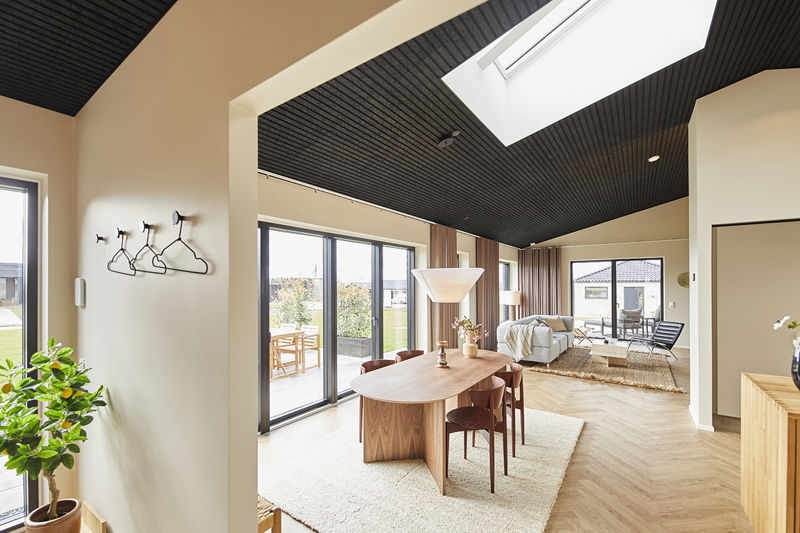 Åbent opholdsrum fra eurodan-huse med spisebord i træ, hvide pendler, store vinduespartier, sort akustikloft med ovenlys, lyse gulve og direkte kig fra køkken-alrum til stue med sofa og loungestol – inspiration til nybyggeri med sammenhæng mellem madlavning, spisning og familieliv.