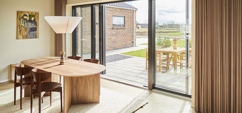 Spiseområde i nybyggeri med sildebensgulv, træbord, gulvlange gardiner og store skydedøre til den overdækkede terrasse.