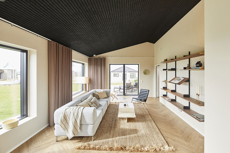 Lang stue fra eurodan-huse med lys sofa langs vinduesvæggen, sort loungestol, hvidt sofabord, stort jutetæppe, åbne træhylder på væggen, brune gardiner og terrassekig – inspiration til nybyggeri, husbyggeri og moderne husdrømme.
