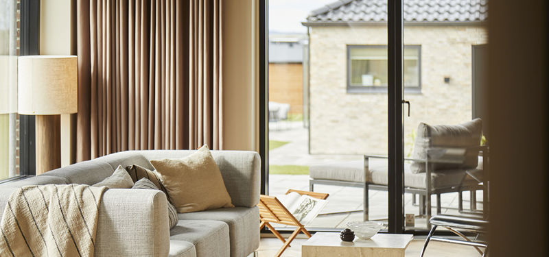 Stue fra eurodan-huse med markante gulv-til-loft-gardiner, stor sofa, lyse trægulve, brede vinduespartier, rolig loungeindretning og mørkt akustikloft – inspiration til nybygget hus, husbyggeri og indretning af stue med varme tekstiler.