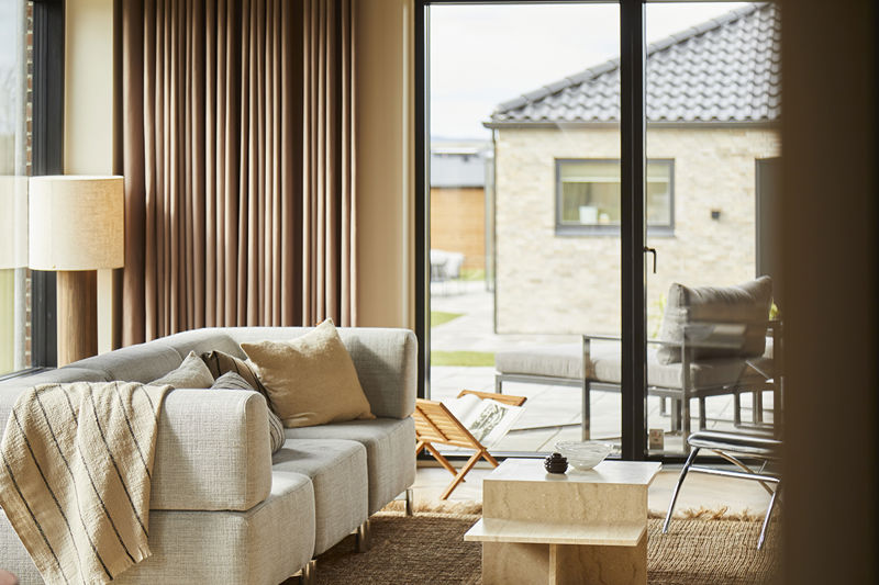 Stue fra eurodan-huse med markante gulv-til-loft-gardiner, stor sofa, lyse trægulve, brede vinduespartier, rolig loungeindretning og mørkt akustikloft – inspiration til nybygget hus, husbyggeri og indretning af stue med varme tekstiler.