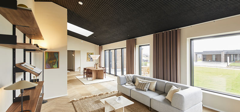 Åben stue fra eurodan-huse med lys modulsofa, hvidt sofabord, stort naturtæppe, sort akustikloft, lange brune gardiner, reolvæg i træ og kig mod spiseplads – inspiration til moderne nybyggeri, husbyggeri og åbne opholdsrum.