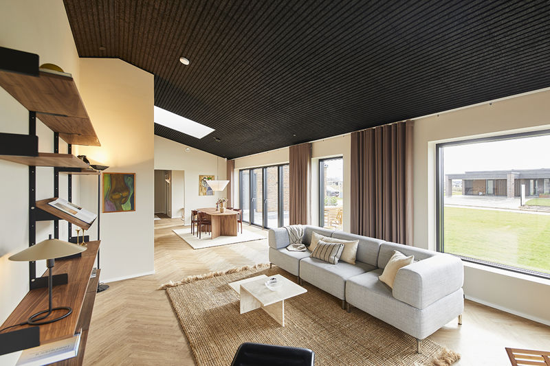Åben stue fra eurodan-huse med lys modulsofa, hvidt sofabord, stort naturtæppe, sort akustikloft, lange brune gardiner, reolvæg i træ og kig mod spiseplads – inspiration til moderne nybyggeri, husbyggeri og åbne opholdsrum.