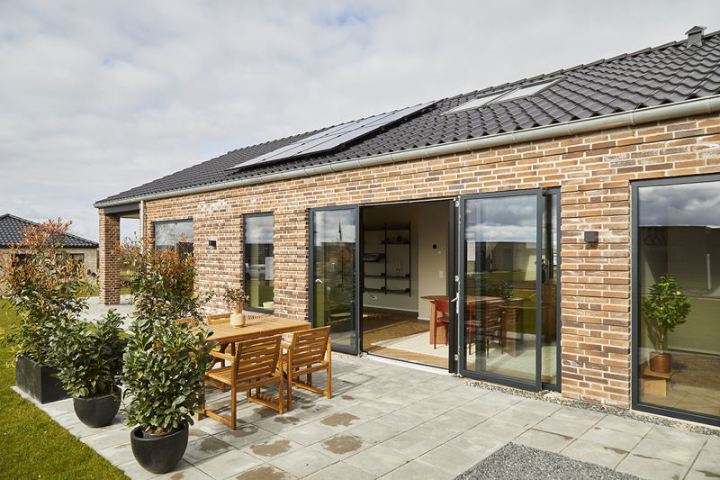 Nærbillede af terrasse fra eurodan-huse med åbne skydedøre i mørke rammer, murstensfacade, træmøbler, plantekrukker og direkte kig ind til opholdsrum – inspiration til nybyggeri med flydende overgang mellem inde og ude.