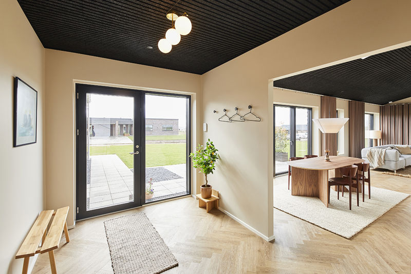 Entré og opholdsrum fra eurodan-huse med sort hoveddør, stort sidevindue mod haven, sildebensgulv, bænk, knagerække og kig videre til spiseplads og stue – inspiration til lys entré, døre og nybyggeri med åbne rum.