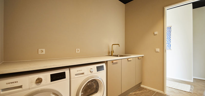 Bryggers fra eurodan-huse med hvid vaskemaskine og tørretumbler under lys bordplade, beige underskabe, lille vask med messingfarvet armatur, mørkt klinkegulv og åben forbindelse til næste rum – inspiration til funktionelt nybyggeri, praktisk husbyggeri og hverdagsvenlige husdrømme.