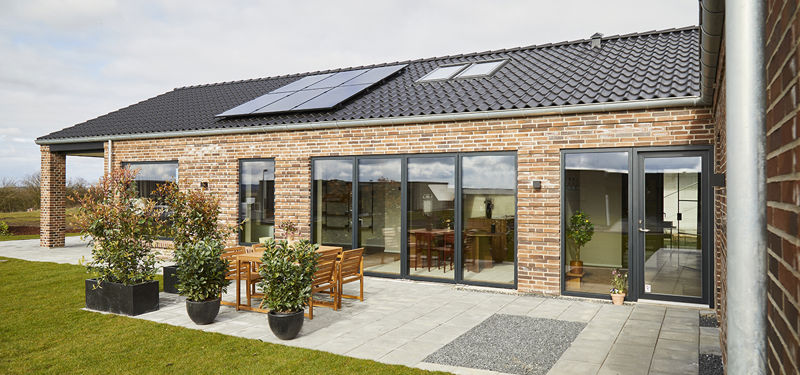 Terrasse foran murstensfacade fra eurodan-huse med store mørke skydedørspartier, træbord med havestole, krukker med grønne planter, lyse terrassefliser, græsplæne og sort tag med solceller – inspiration til nybyggeri, uderum og moderne husdrømme.