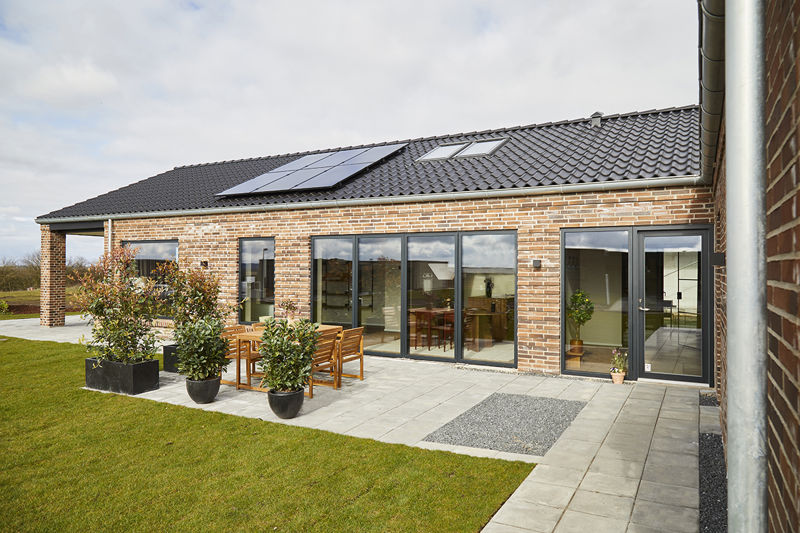 Terrasse foran murstensfacade fra eurodan-huse med store mørke skydedørspartier, træbord med havestole, krukker med grønne planter, lyse terrassefliser, græsplæne og sort tag med solceller – inspiration til nybyggeri, uderum og moderne husdrømme.