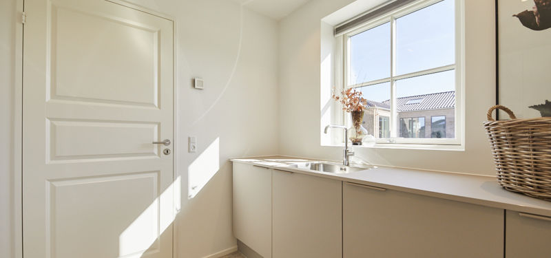 Lyst bryggers fra eurodan-huse med lang bordplade, underskabe i lyse fronter, integreret vask under stort vindue, hvid fyldningsdør og dekorativ vase ved vindueskarmen – inspiration til nybyggeri med funktionelt bryggers, god opbevaring og lyse husdrømme.