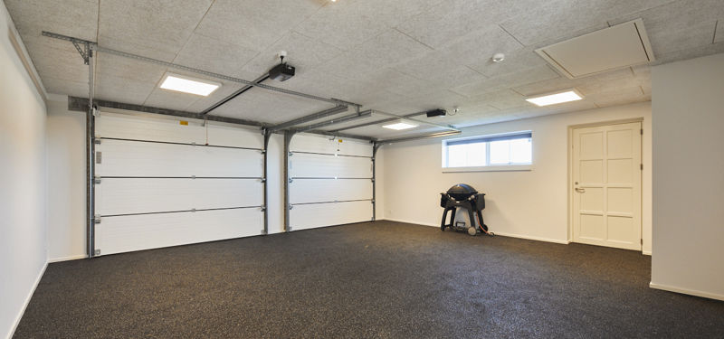 Indvendig dobbelt garage med to hvide garageporte, sort gulv, loftslamper, lille vindue, hvid dør og god gulvplads til biler eller opbevaring – inspiration til garage i nybyggeri og funktionelle husdrømme fra eurodan-huse.