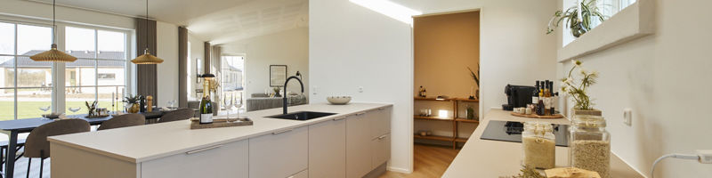 Køkken fra eurodan-huse med lys køkkenø, sort vask og armatur, lang bordplade langs vinduet med glas, krukker og planter, smalt højtsiddende vindue, synligt depotrum med hylder, stort ovenlysvindue og spiseplads i siden – inspiration til praktisk nybyggeri, husbyggeri og køkkendrømme.