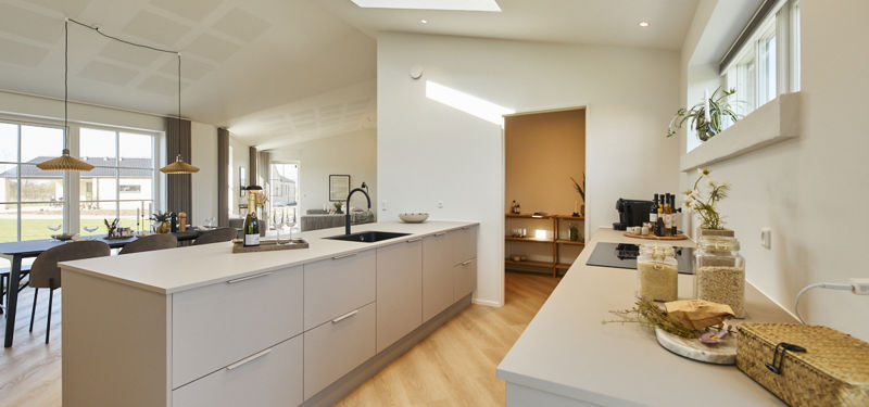 Køkken fra eurodan-huse med lys køkkenø, sort vask og armatur, lang bordplade langs vinduet med glas, krukker og planter, smalt højtsiddende vindue, synligt depotrum med hylder, stort ovenlysvindue og spiseplads i siden – inspiration til praktisk nybyggeri, husbyggeri og køkkendrømme.