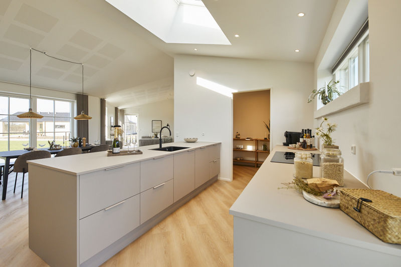 Køkken fra eurodan-huse med lys køkkenø, sort vask og armatur, lang bordplade langs vinduet med glas, krukker og planter, smalt højtsiddende vindue, synligt depotrum med hylder, stort ovenlysvindue og spiseplads i siden – inspiration til praktisk nybyggeri, husbyggeri og køkkendrømme.