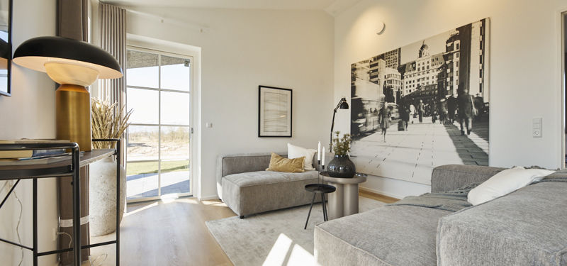 Stue fra eurodan-huse med to grå loungemoduler, rundt sidebord, sort standerlampe, stor sort-hvid vægkunst, lys gulvflade og dobbelte vinduer mod terrasse – inspiration til stilren stue i nybyggeri, husbyggeri og nybygget hus.