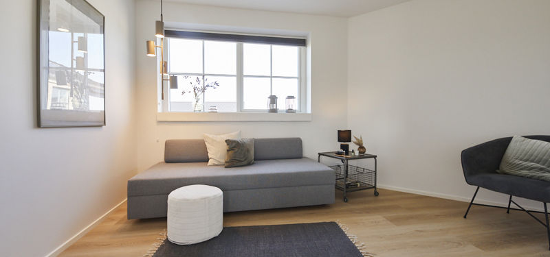 Lyst og fleksibelt værelse med grå sovesofa, mørk loungestol, lille sidebord, gulvlampe og bredt sprossevindue – inspiration til gæsteværelse eller opholdsrum i nybyg.