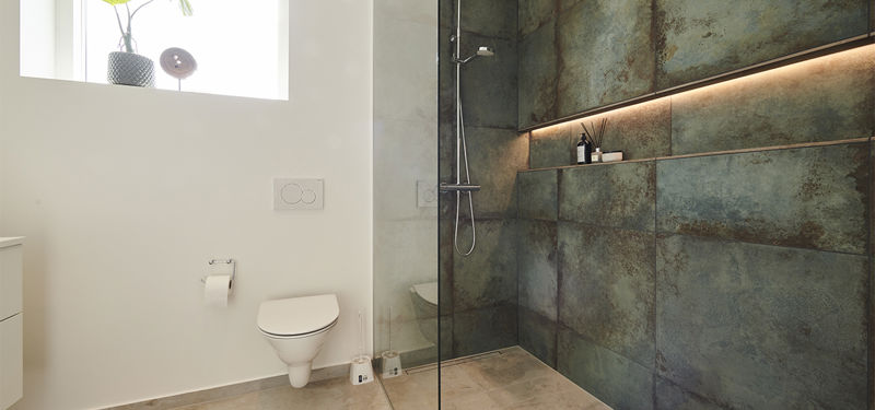 Badeværelse fra eurodan-huse med væghængt toilet, højtsiddende vindue, glasvæg til bruseniche, mørke fliser med råt stenlook, indbygget shampoohylde med lys og synligt brusesæt – inspiration til moderne nybyggeri, husbyggeri og funktionelle badeværelsesløsninger.