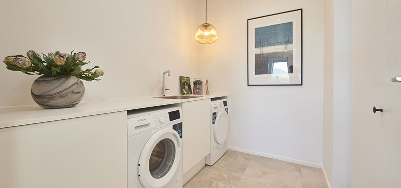 Bryggers fra eurodan-huse med to hvide vaskemaskiner under lang bordplade, indbygget vask, marmorinspireret vase med blomster, rund pendel og grafisk vægkunst – inspiration til nybyggeri med praktisk vaskeplads, funktionelt husbyggeri og moderne husdrømme.