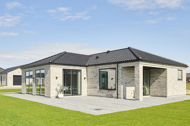 Fliseterrasse fra eurodan-huse rundt om lys murstensfacade med sort tegltag, store glasdøre, bredt vindue, overdækket terrassehjørne og grøn græsplæne omkring huset – inspiration til nybygget hus med terrasse og uderum.