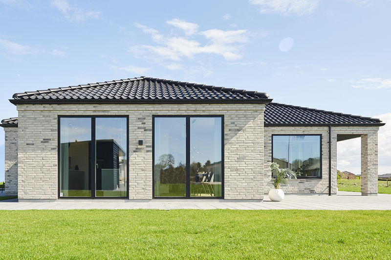 Murstensfacade fra eurodan-huse med sort tegltag, brede vinduespartier, mørke terrassedøre, flisebelagt terrasse og stor græsplæne foran huset – inspiration til vinduer, døre og klassisk nybyggeri med moderne detaljer.