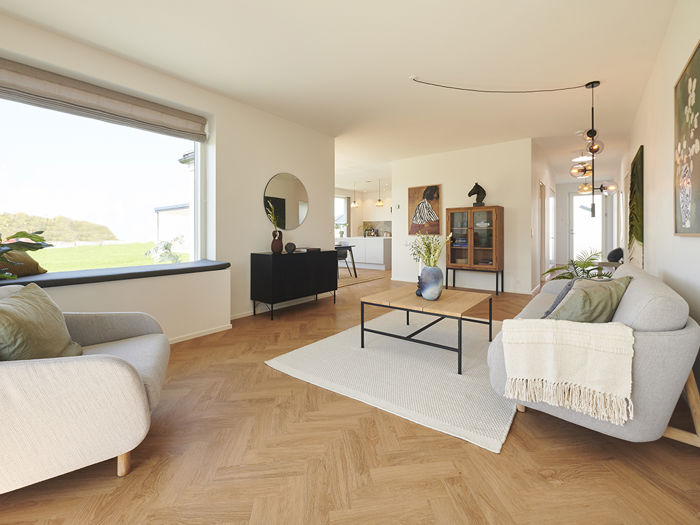 Stue fra eurodan-huse med lyst sildebensgulv, grå sofa, beige lænestol, stort sansevindue med hynde, sort skænk, rundt spejl og åben forbindelse til køkken-alrum – inspiration til funktionelt nybyggeri, husbyggeri og hyggelige husdrømme.