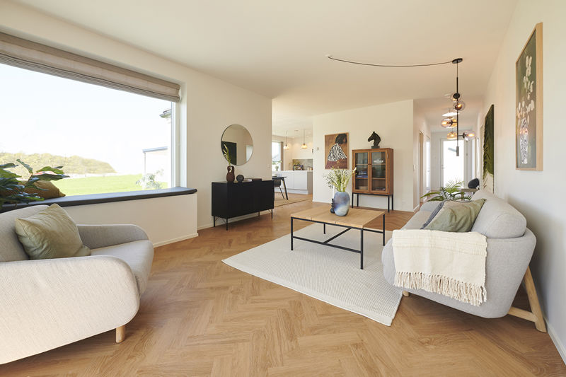 Stue fra eurodan-huse med lyst sildebensgulv, grå sofa, beige lænestol, stort sansevindue med hynde, sort skænk, rundt spejl og åben forbindelse til køkken-alrum – inspiration til funktionelt nybyggeri, husbyggeri og hyggelige husdrømme.