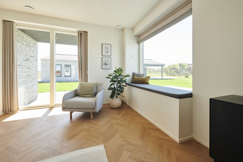 Stuehjørne fra eurodan-huse med beige lænestol, højt terrassevindue med gardiner, stort sansevindue med mørk hynde, grøn plante og sort skænk i hjørnet – konkret inspiration til funktionelt nybyggeri, husbyggeri og moderne husdrømme.