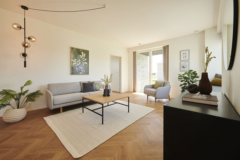 Stue fra eurodan-huse med grå sofa, beige lænestol, lyst tæppe, træbord, sort pendel med flere glaskupler, stor plante og bred udgang mod overdækket terrasse – inspiration til nybyggeri, husbyggeri og husdrømme med lys stue.