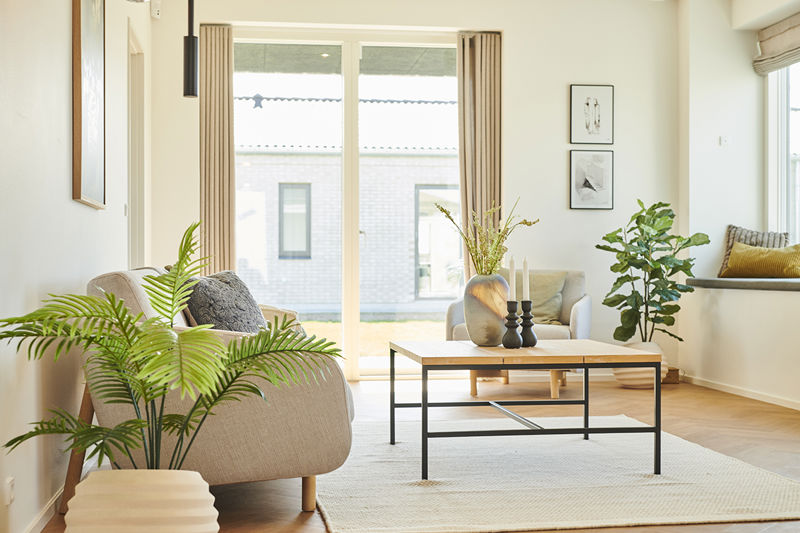 Stue fra eurodan-huse med sofaområde, beige lænestol, firkantet sofabord, store terrassevinduer med gardiner, grønne planter og sansevindue med hynde i siden – inspiration til nybyggeri, husbyggeri og indretning af nybygget hus.