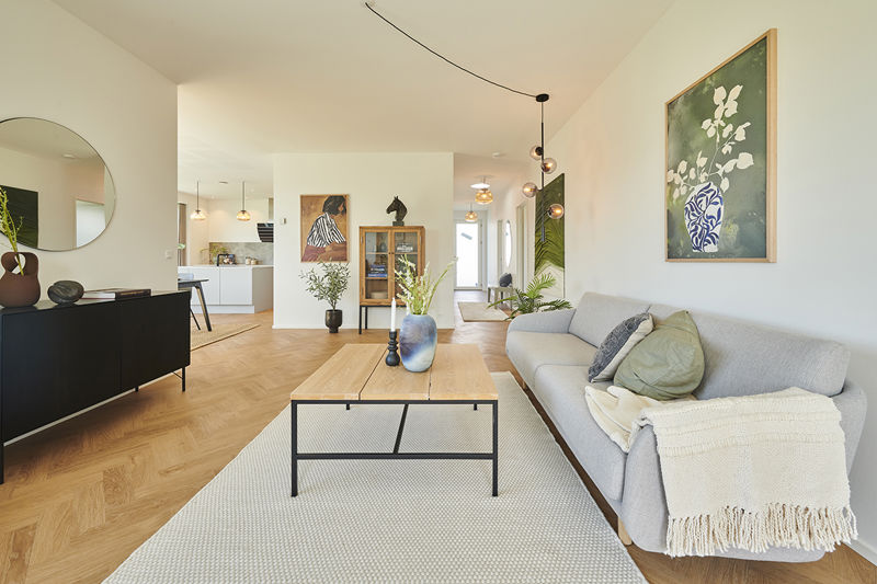 Stue fra eurodan-huse med lysegrå sofa, grønne puder, lyst tæppe, sofabord i træ og sort metal, sort skænk, stor rund spejl og åben forbindelse til hvidt køkken – inspiration til moderne nybyggeri, husbyggeri og husdrømme.