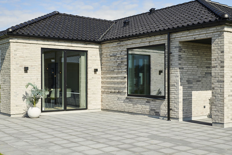 Stor fliseterrasse fra eurodan-huse med lyse mursten, sorte glasdøre, sort tegltag, overdækket hjørne, væglamper og krukkeplante ved facaden – inspiration til terrasse i nybyggeri og moderne husbyggeri med gode uderum.