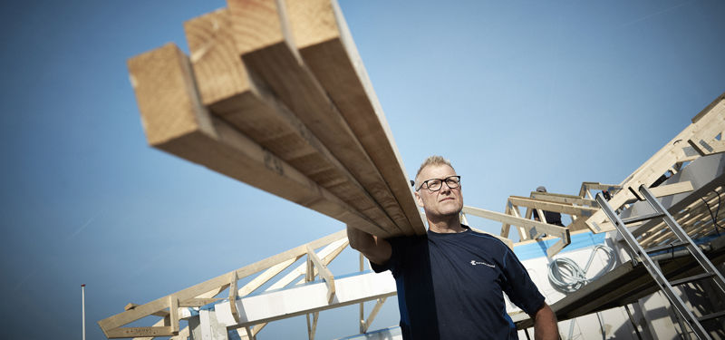 Håndværker bærer træbjælker på byggepladsen foran tagkonstruktionen til et nyt hus under opførelse.
