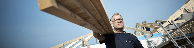 Håndværker bærer træbjælker på byggepladsen foran tagkonstruktionen til et nyt hus under opførelse.